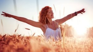Woman standing in a sunlit grain field with arms outstretched, symbolizing freedom, self-liberation, and reclaiming one’s natural rhythm beyond societal expectations.