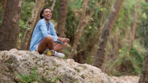 Woman sitting on a rock in nature, smiling with peaceful presence, symbolizing recovery, resilience, and connection to body wisdom.