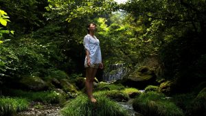 Woman standing alone in a forest, gazing upward with a sense of openness and reflection, symbolizing personal reclamation and energetic renewal.