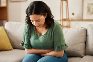 Woman sitting on a couch with arms over her belly, looking down with concern — symbolizing the emotional and physical discomfort of menopause-related anxiety and gut distress.