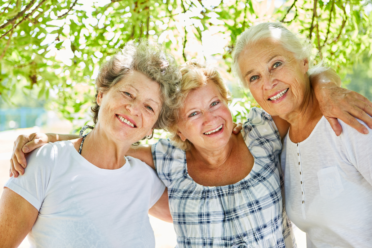 Menopause and elder wisdom: why your presence matters now Three women standing with arms around each other, smiling with warmth and connection — symbolizing sisterhood, elder wisdom, and the power of post-menopausal presence.