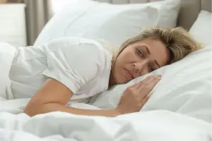 Woman lying on her side in white bed sheets, staring awake into the distance — representing sleep disruption during menopause and the deeper causes behind it.
