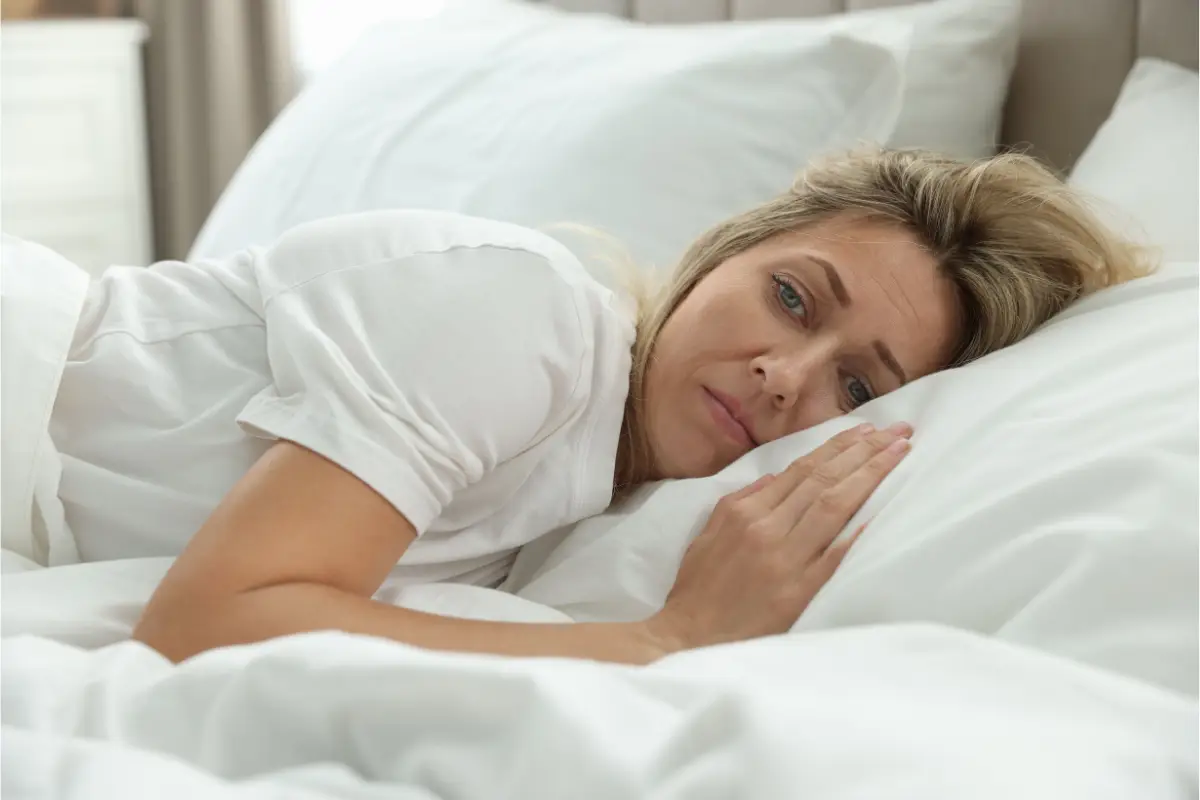 Sleep and menopause: why it’s not just hormones Woman lying on her side in white bed sheets, staring awake into the distance — representing sleep disruption during menopause and the deeper causes behind it.